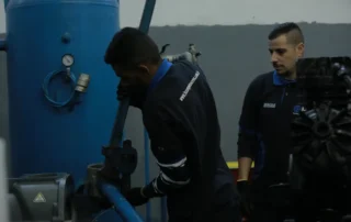 Técnicos realizando manutenção em sistema de compressor industrial e reservatório de ar comprimido em ambiente de fábrica.