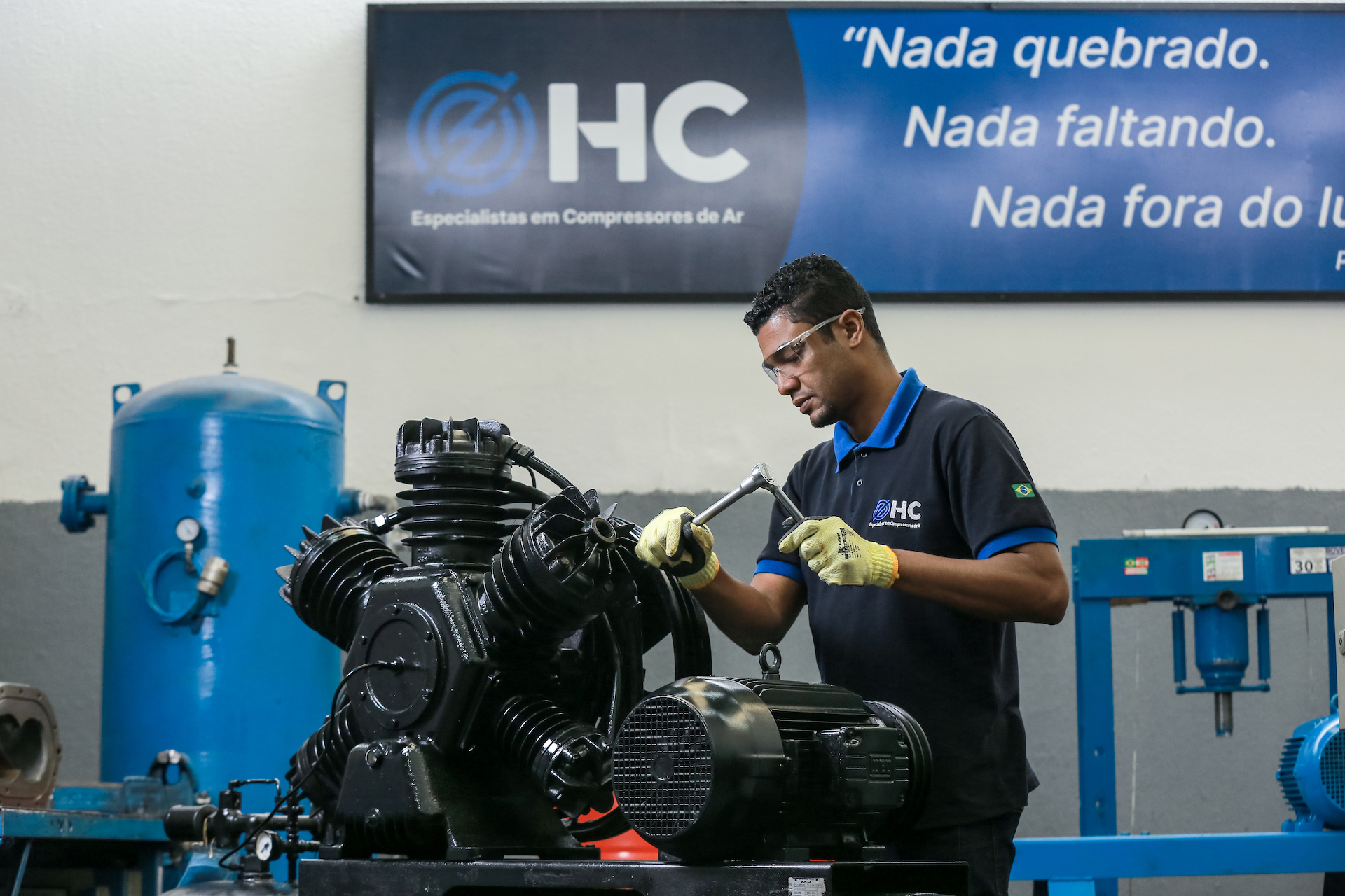 Técnico da HC Compressores montando o cabeçote de um compressor de pistão industrial em bancada de oficina, sob o painel com o lema da empresa.