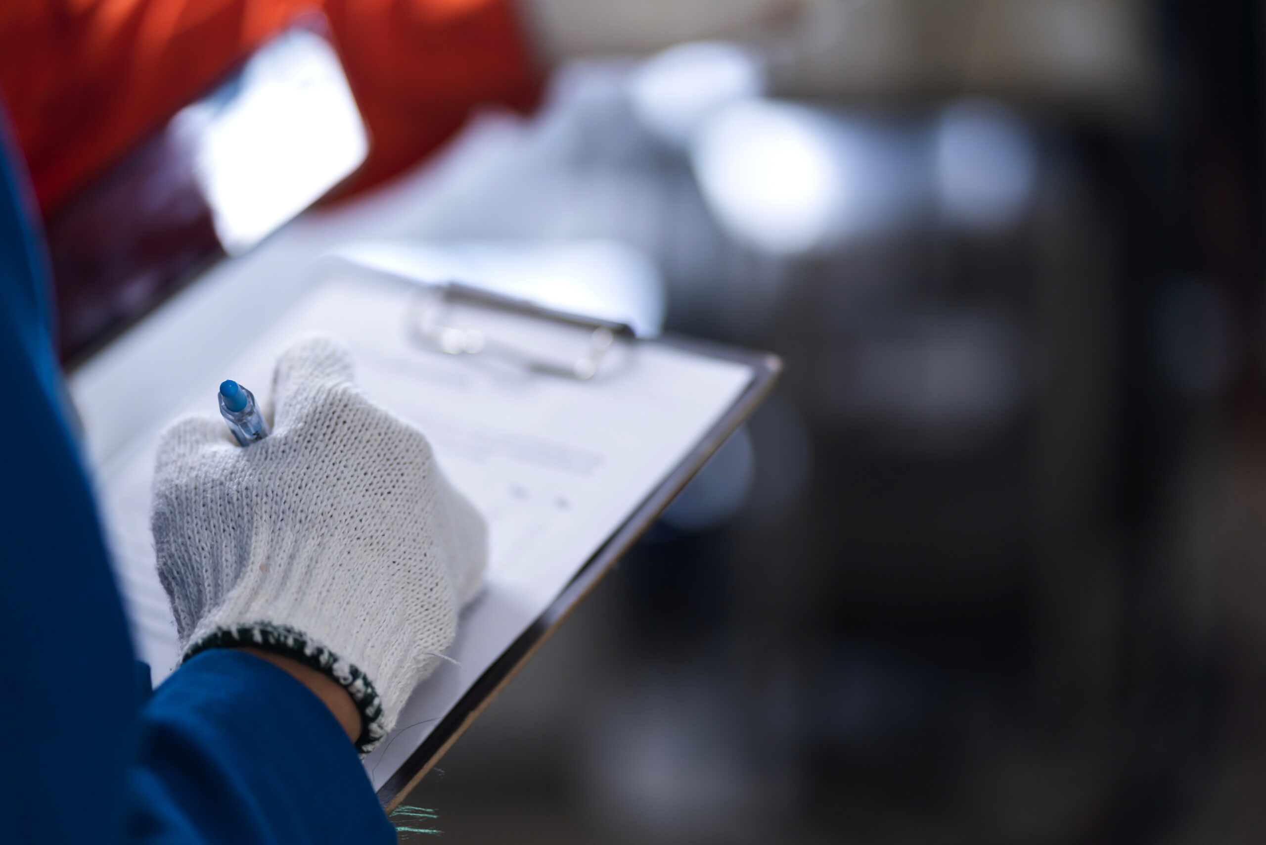 Mão com luva de proteção preenchendo formulário de inspeção em prancheta, com equipamentos industriais desfocados ao fundo.