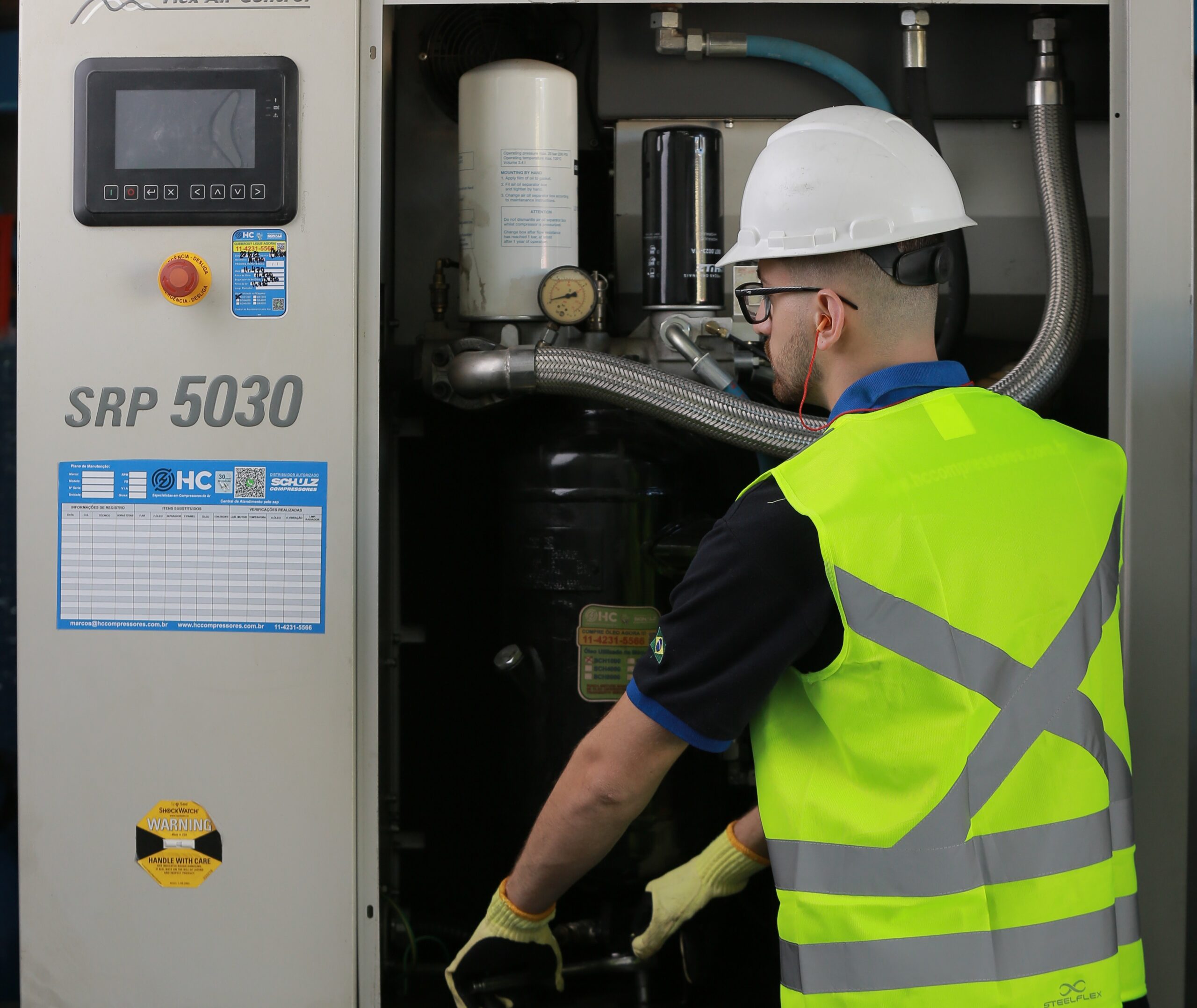Técnico da HC Compressores em manutenção de compressor de parafuso Schulz SRP 5030, com plano de manutenção afixado no painel e componentes internos expostos.