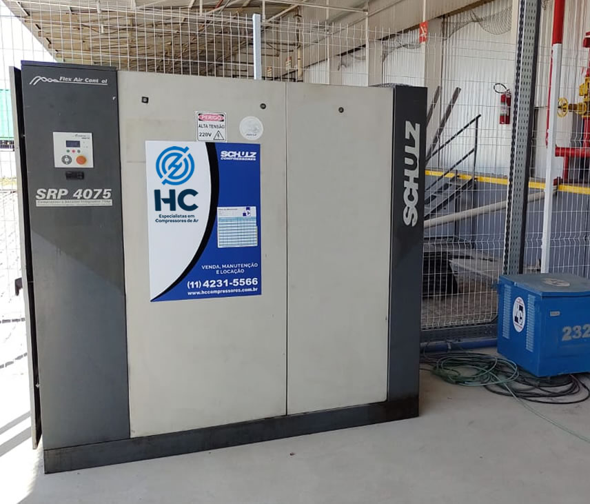 Compressor de parafuso Schulz SRP 4075 instalado em área externa cercada, com etiqueta de identificação e contato da HC Compressores no painel frontal.