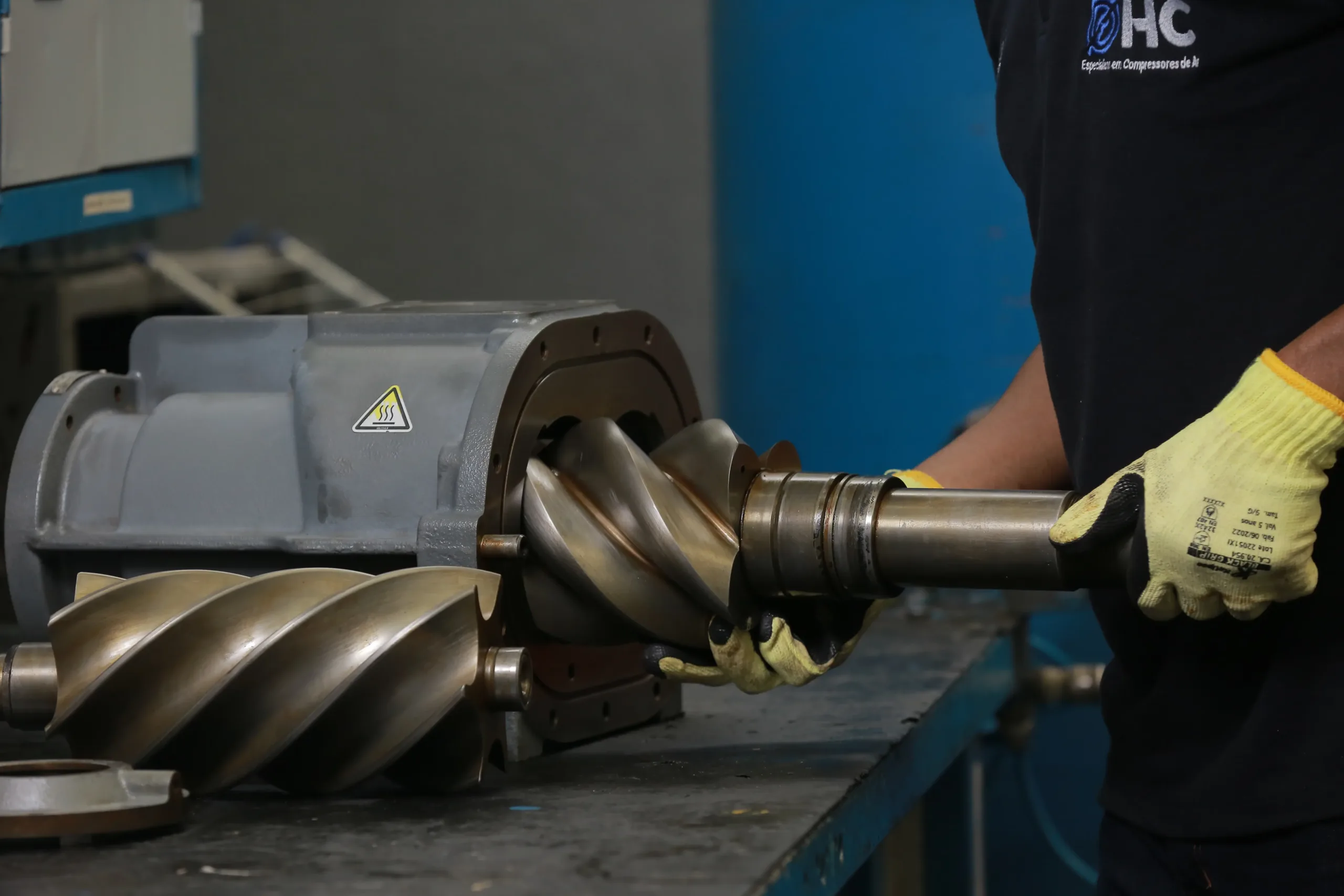 Técnico da HC Compressores com luvas de proteção removendo o par de rotores helicoidais de uma unidade compressora de parafuso sobre bancada de oficina.