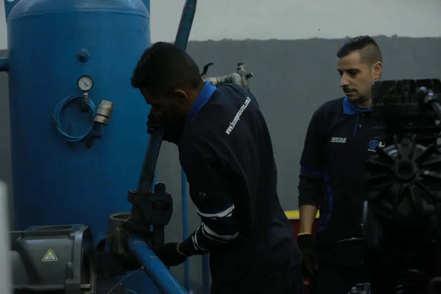 Técnicos realizando manutenção em sistema de compressor industrial e reservatório de ar comprimido em ambiente de fábrica.