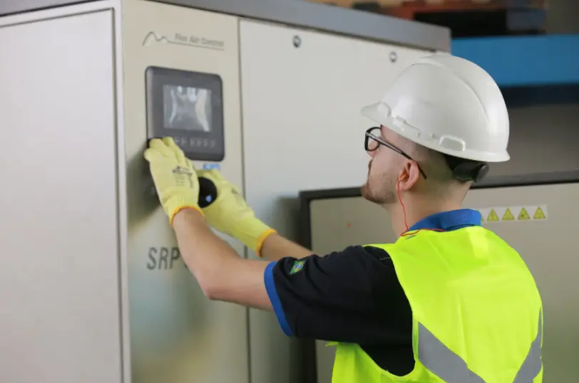 Técnico com capacete e luvas de proteção ajustando os parâmetros de pressão no painel eletrônico de um compressor de parafuso Schulz SRP.