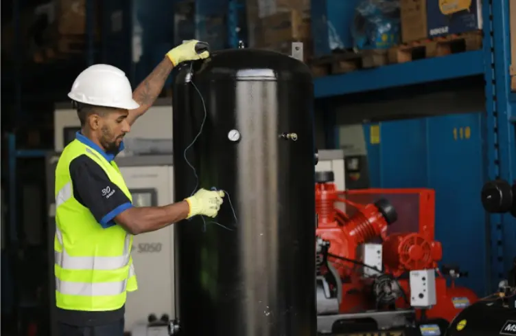Técnico com capacete e colete refletivo inspecionando um reservatório vertical de ar comprimido em oficina, com compressor de pistão vermelho ao fundo.