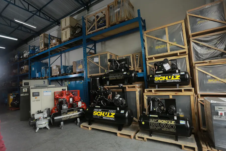 Compressor de ar Schulz de diferentes modelos expostos em estoque industrial, com equipamentos sobre pallets e estruturas metálicas.