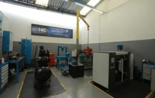 Sala industrial da HC Compressores com diversos equipamentos de ar comprimido, incluindo compressores e reservatórios.