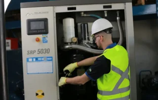 Técnico realiza manutenção programada em compressor de ar industrial, inspecionando componentes internos do equipamento com uso de EPIs em ambiente fabril.